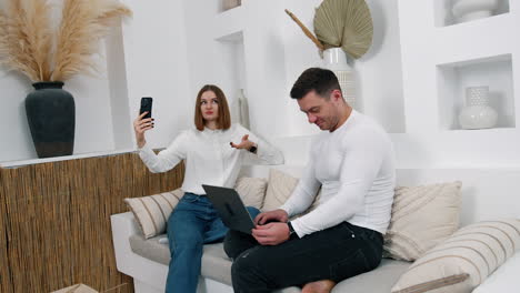 woman holding a phone taking selfie of having video chat. man sitting beside is focused on his laptop.