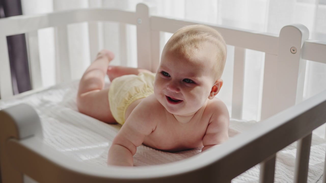 Infant with blonde hair wearing yellow shorts lies on stomach in white crib looking down with a