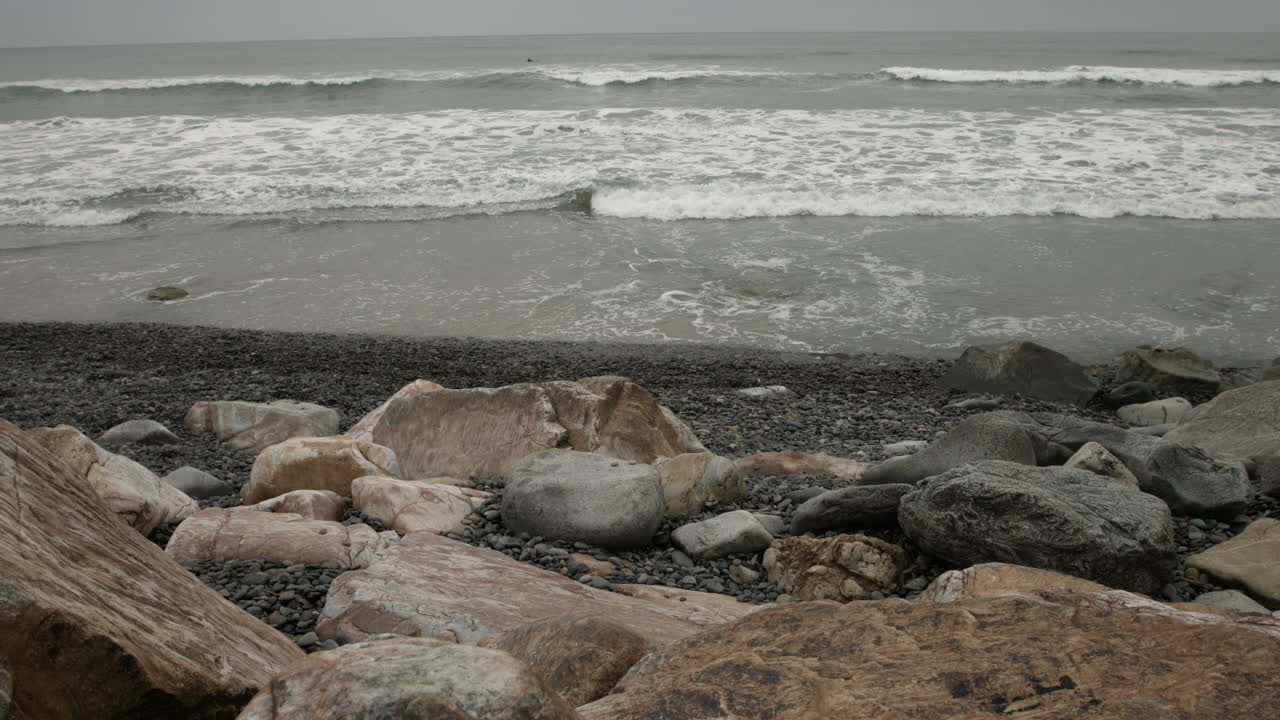 A UHD wide-angle shot captures a quiet rocky beach on an overcast morning, with soft gray light, textured stones, and gentle waves creating a serene, moody, and contemplative coastal scene.