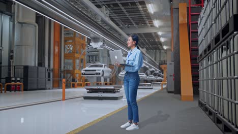 vista lateral completa del cuerpo de una trabajadora asiática de pie en una fábrica de automóviles ev, instalación industrial de alta tecnología, observa mirando hacia arriba antes de que ella venga a concentrarse en la computadora portátil y seguir escribiendo