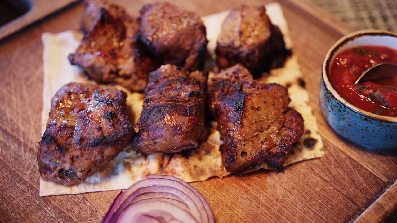 Pezzi di carne grigliata sono disposti su pane piatto, serviti con una piccola ciotola di salsa rossa e fette di cipolla rossa su una superficie di legno.