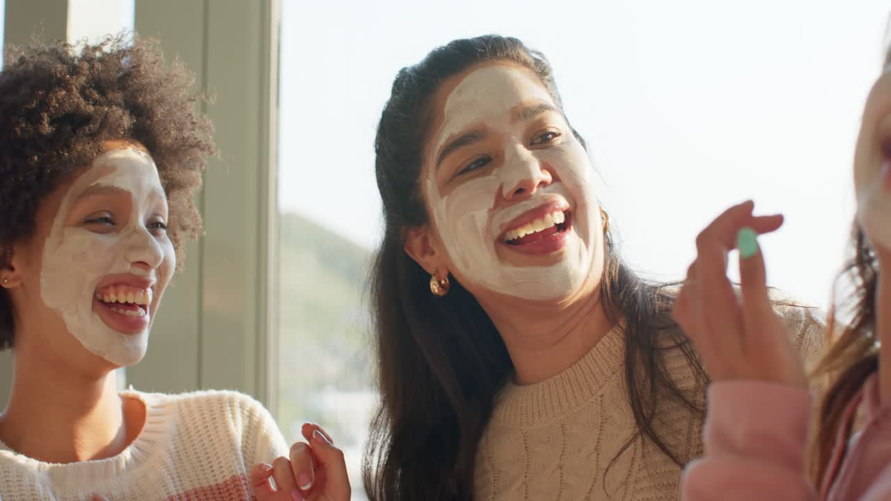 Diverse female friends with masks tapping cheeks laughing, third entering adjusting mask in lounge
