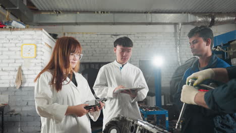 engineer demonstrates loosening bolt while students take notes on notepads, another colleague observes attentively in an automotive workshop filled with tools, and car engines in background