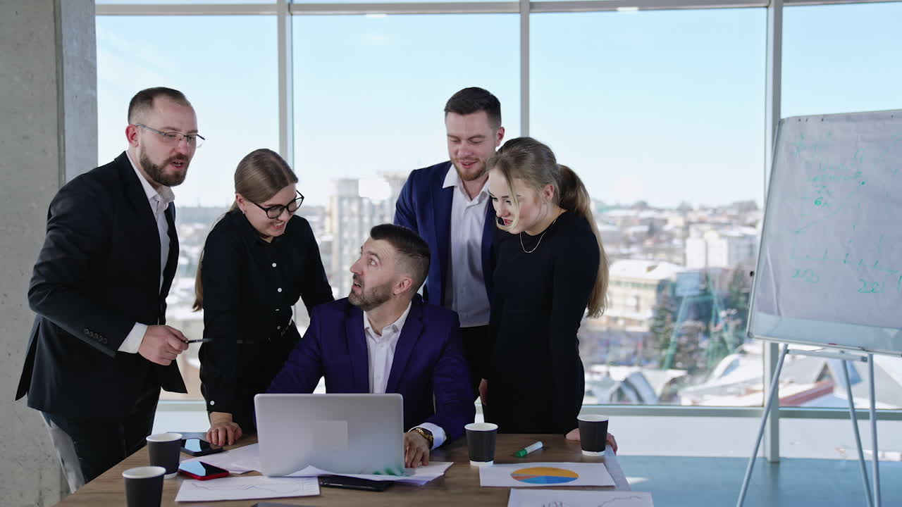 Boss sitting in front of laptop and other coworkers stand beside him. Brainstorming and discussing ideas in the positive and easy working atmosphere. Young collective solving working tasks.