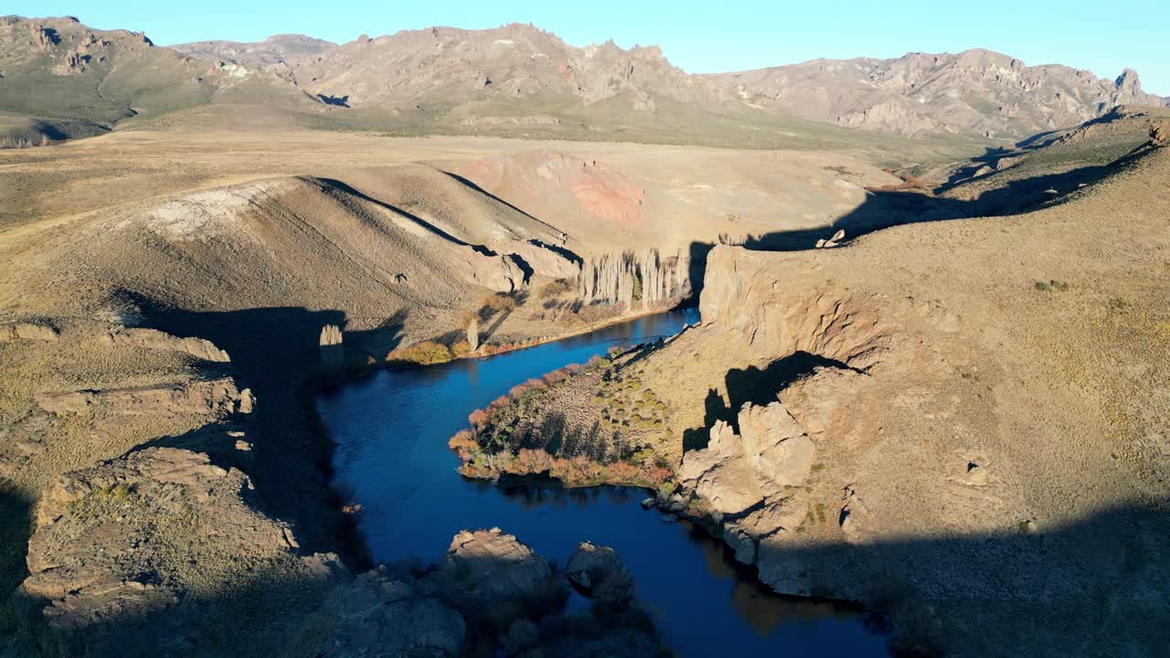Sweeping aerial footage of a river near Bariloche, Argentina, surrounded by golden fields and green slopes at sunset
