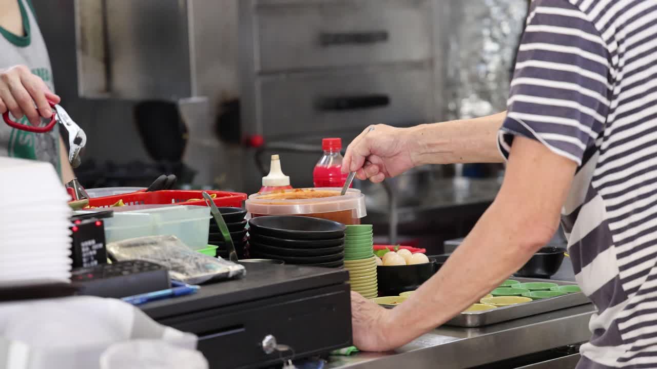 Two vendors assemble meals at a hawker center counter under bright, even indoor lighting