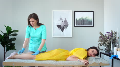 two girls in a beauty salon master in a bathrobe and gloves doing the procedure to remove hair on the legs with a sugar mixture