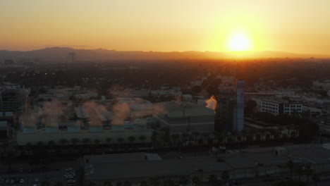 Antena:-Fábrica-Humeante-Con-Palmeras-Y-Carretera-Transitada-En-Burbank,-Los-ángeles,-California,-Puesta-De-Sol