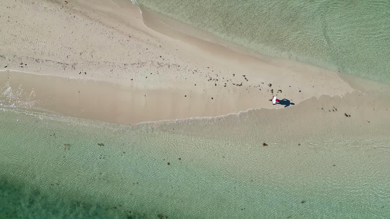 Drone overhead reveals and follows one person walking under the sun on tropical sandbar beach as gentle Lagoon waves lapping on the sides