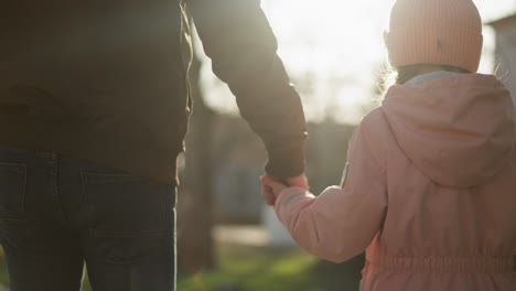 back view of a little girl in a pink cap and jacket, and a man in a brown jacket, both wearing blue jeans, holding hands while walking on a sunny street, with a warm