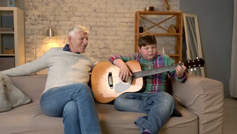 the grandson plays for his grandfather guitar, music. an elderly man is sitting on with a young fat guy playing guitar. home comfort, family idyll, cosiness concept, difference of generations. 60 fps