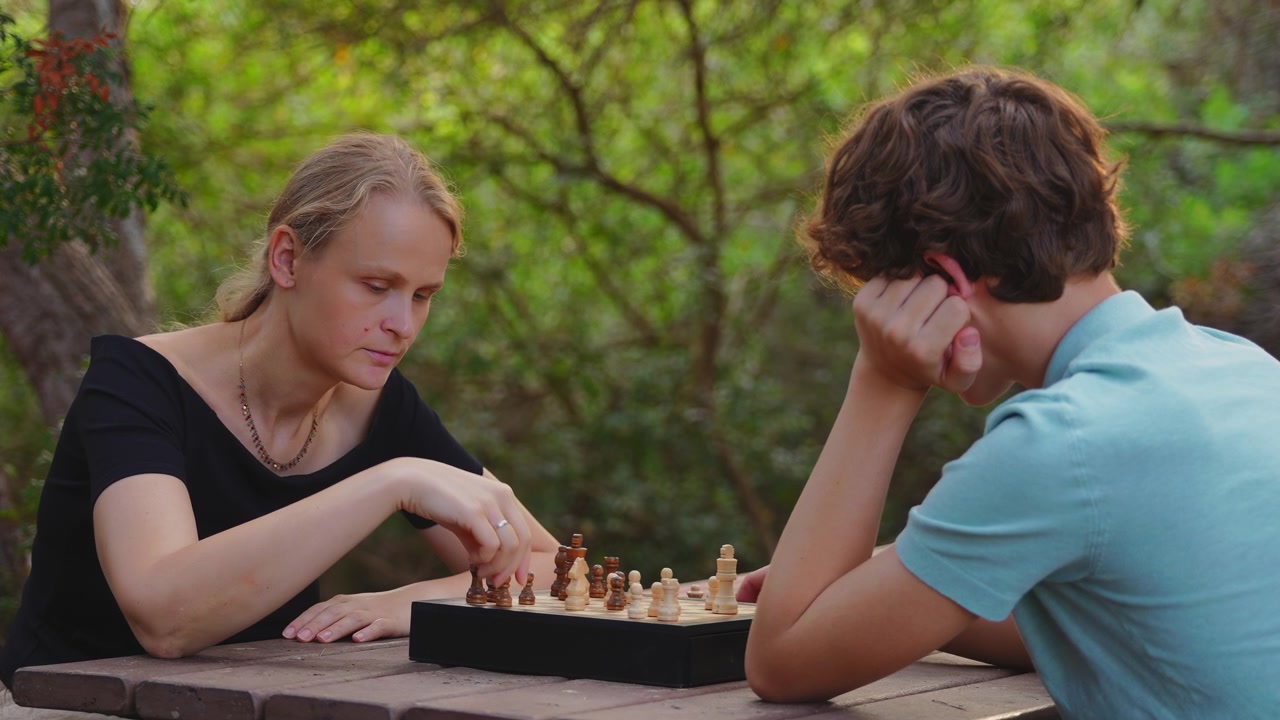 Eine Mutter und ihr Teenager-Sohn sitzen an einem hölzernen Picknicktisch in einem Park und konzentrieren sich auf ein Schachspiel, wobei sie abwechselnd Holzfiguren auf einem von grünem Laub umgebenen Brett bewegen.