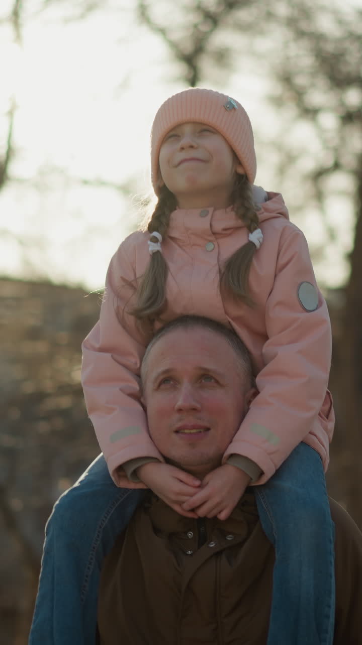 vista anteriore di una ragazzina con un berretto e una giacca rosa seduta gioiosamente sul collo di suo padre mentre camminano attraverso un parco illuminato dal sole. la ragazza sorride con gli occhi aperti, godendosi il momento