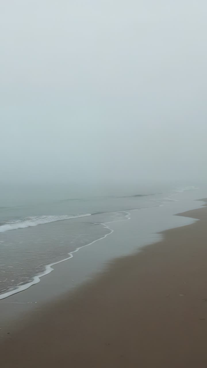Vertical video: Advancing wave crests lapping sandy beach at foggy shore, leaving thin foam line