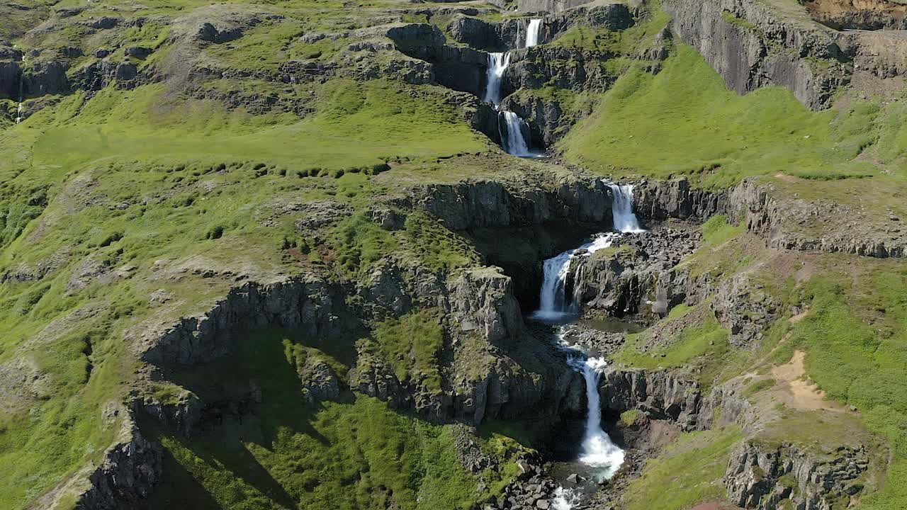 Beautiful aerial dolly in on foamy waterfall running over several ledges framed by mossy green slopes. Kilfbrekkufossar, Icelandic waterfall.