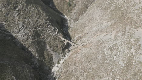 Drone-shot-of-the-Ali-Pasha-Bridge-Outside-of-Gjirokaster,-Albania-on-a-sunny-day-sitting-in-between-the-rocky-mountains-with-a-dry-river-LOG