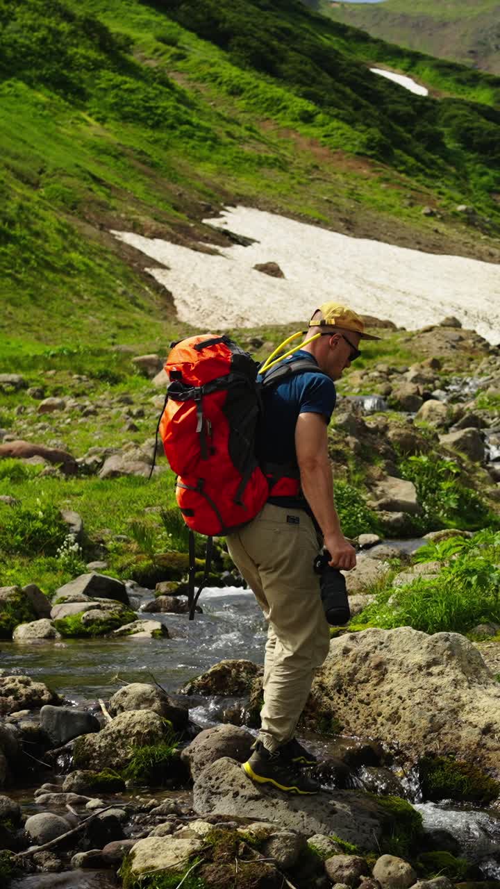 Hombre con mochila y cámara haciendo senderismo cruzando un arroyo rocoso en las montañas.