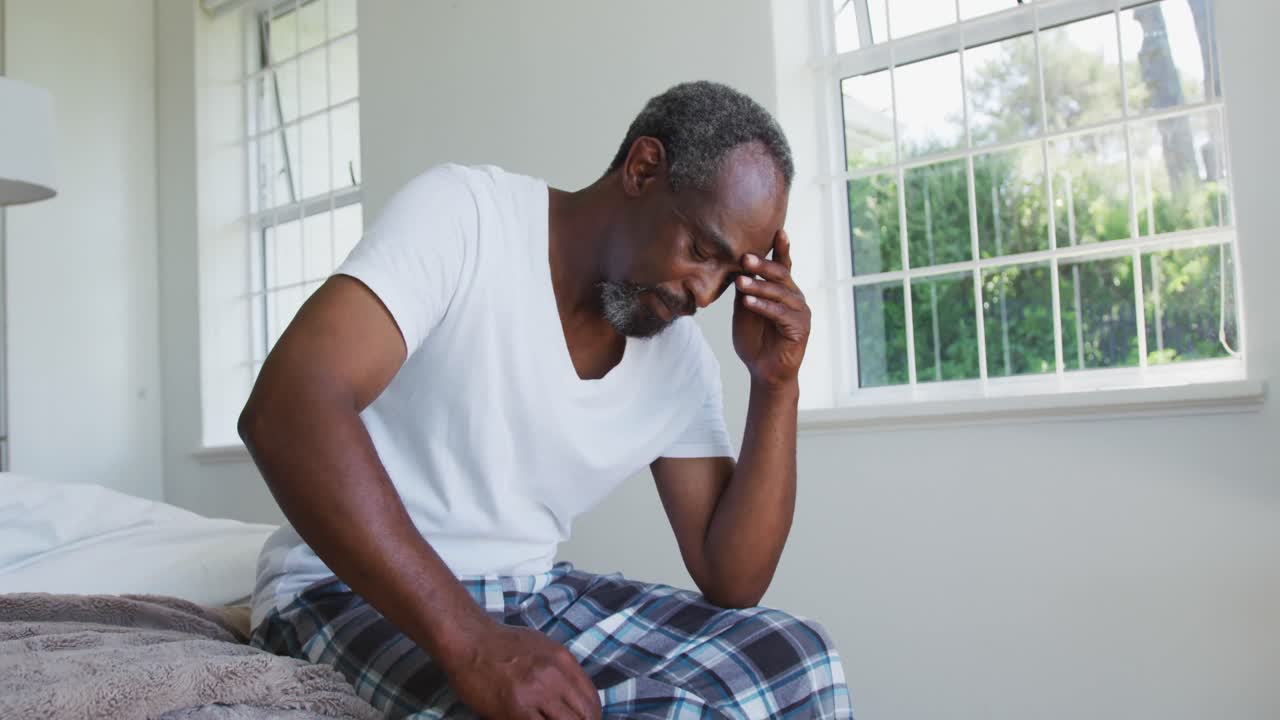 Senior african american man with headache sitting on bed holding head