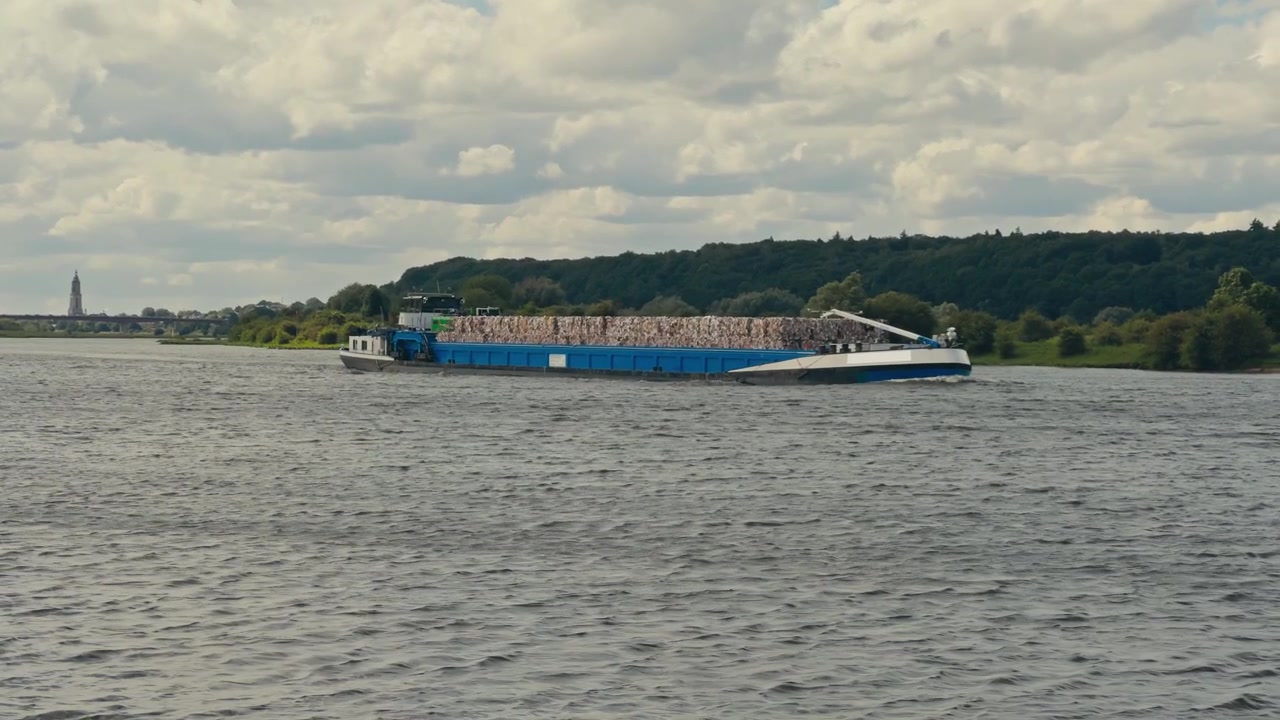 Slowmotion følgeskud af en containerpram, der transporterer gods langs en kanal, med grønne bakker og bløde bomuldsskyer, der skaber en naturskøn sommerbaggrund