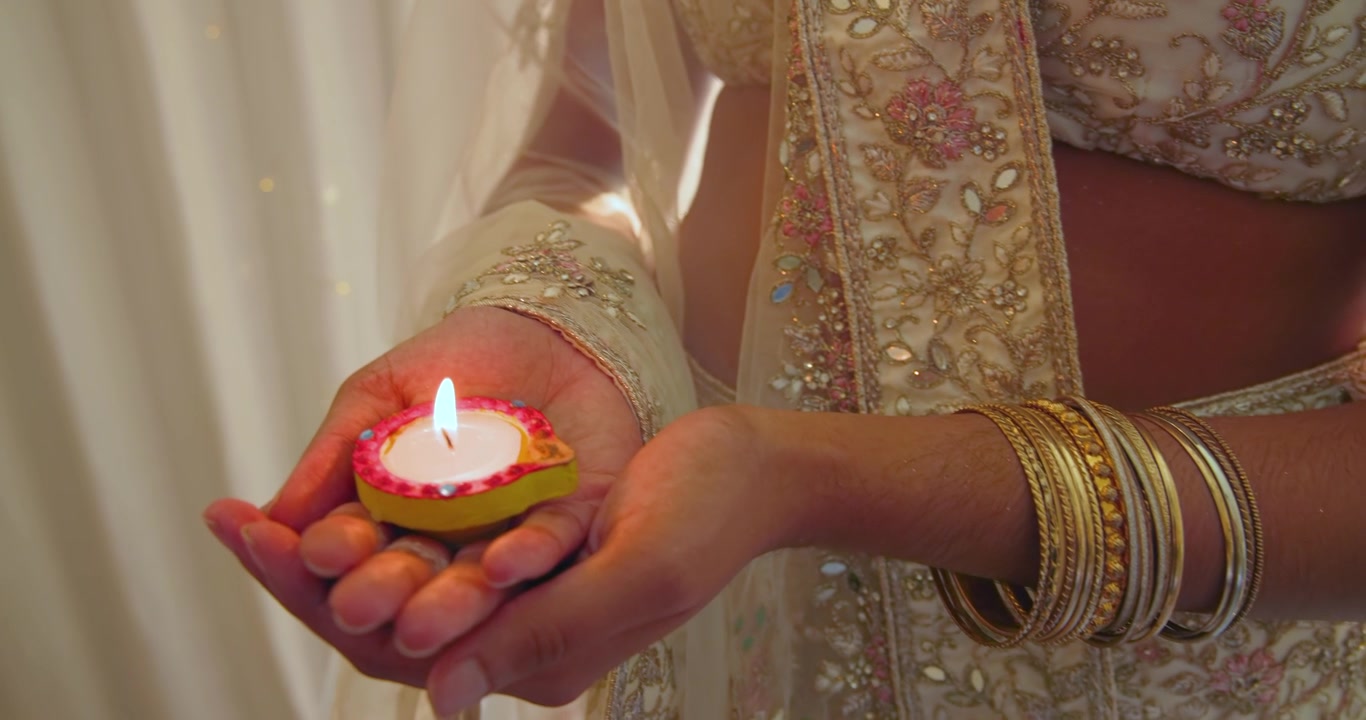 Adult woman cupping lit diya with camera tightening steadying flame showing bangles for ritual