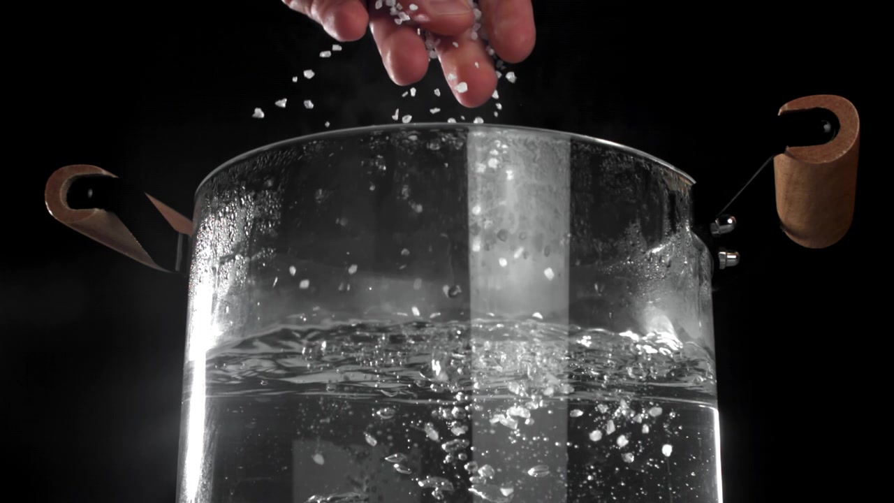 Mano che versa il sale nell'acqua bollente in una pentola di vetro