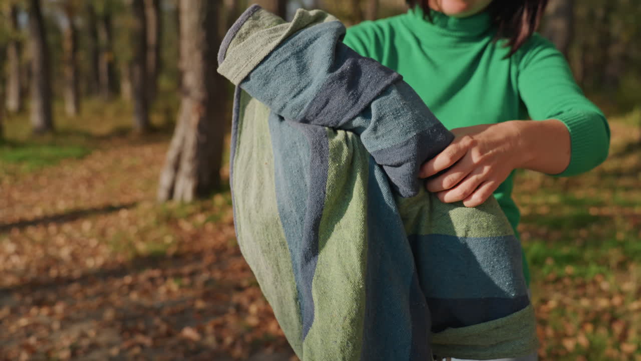 Mujer enrollando una hamaca con cuerda en el bosque, envolviendo la bobina y atando lazos con cordón, formando un paquete compacto para acampar, revisando correas y agarre, bajo la luz solar moteada de otoño, un embalaje eficiente para exteriores.