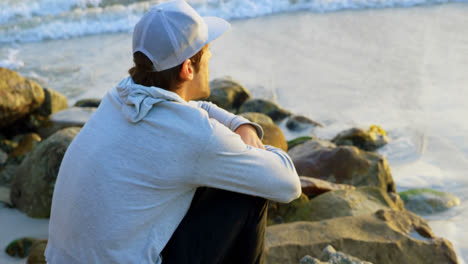 Side-view-of-young-caucasian-man-sitting-on-a-rock-and-looking-at-sea-on-the-beach-4k