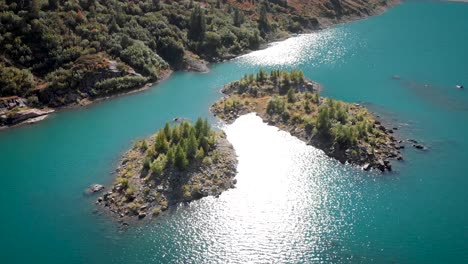 Aerial-flyover-over-sunlit-forested-islands-in-the-turquoise-waters-of-Lac-de-Salanfe-in-Valais,-Switzerland-on-a-sunny-autumn-day-in-the-Swiss-Alps-with-a-pan-down-view