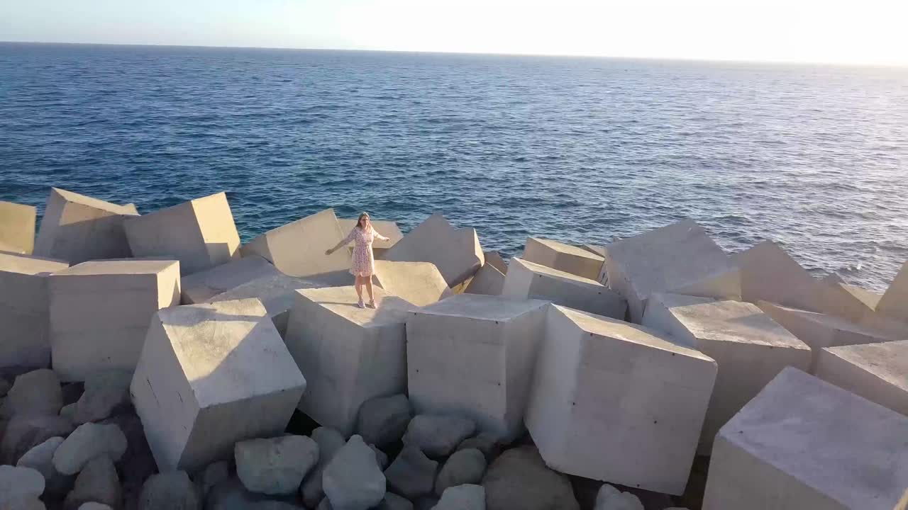 Aerial circle shot of woman standing on rocks at shore during sunny day with raising arms.