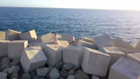aerial circle shot of woman standing on rocks at shore during sunny day with raising arms.