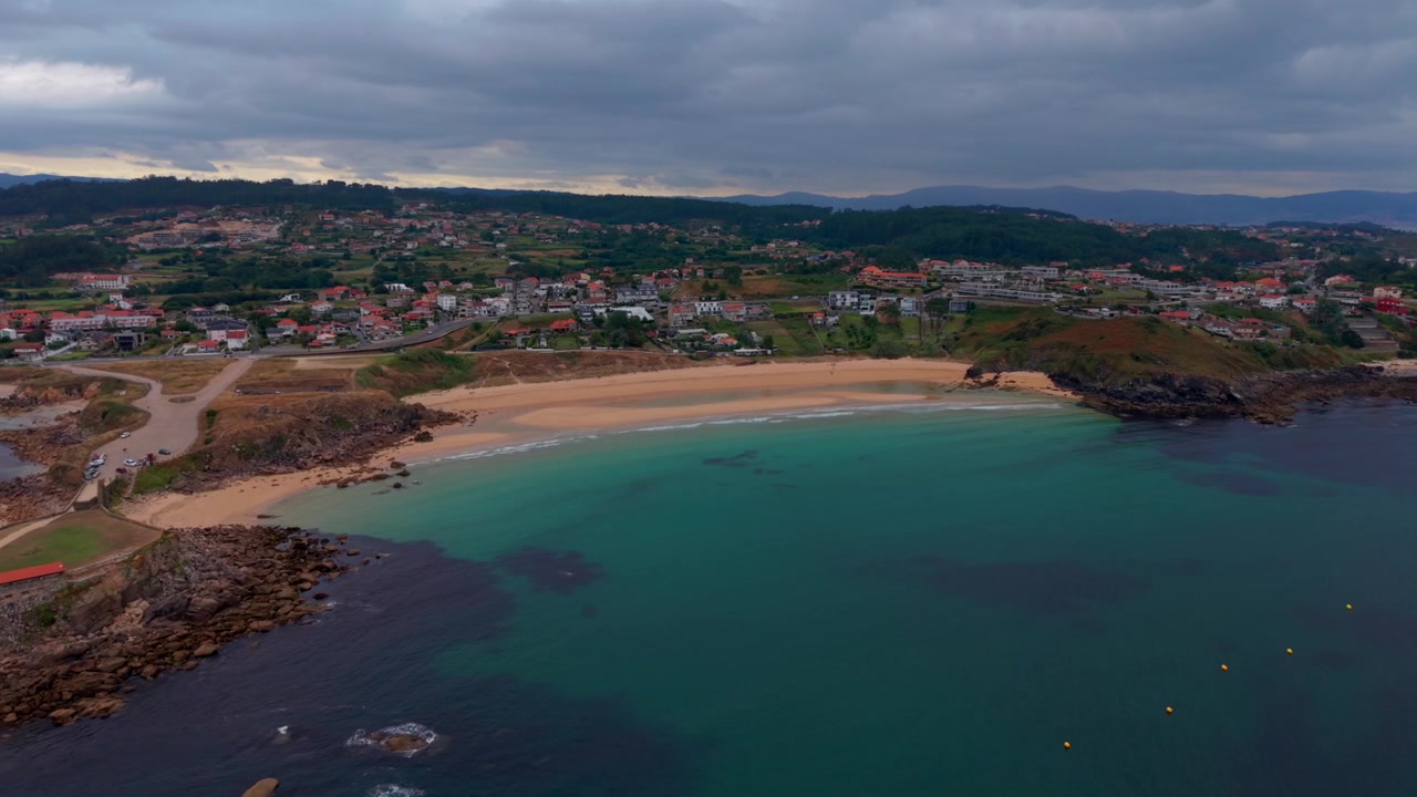 Luftdronebillede af en buet sandstrand i en beskyttet bugt med turkisblåt vand, klippefyldte forbjerge og en kystby i Pontevedra, Galicien, Spanien.