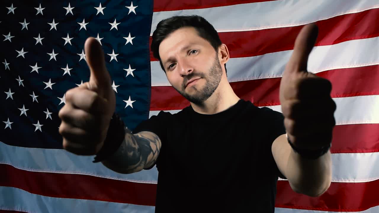 A proud patriot with tattoos, short hair and a beard does thumbs up expression by lifting his arms up and is happy in front of an American United States flag that is slowly moving in the background