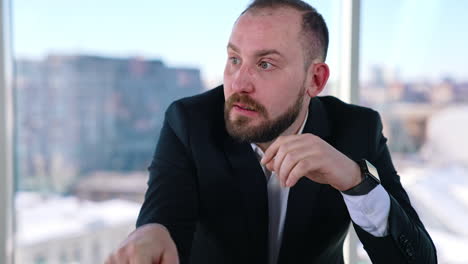 portrait of a tired businessman at workplace. bearded office worker in suit sitting at the table with a laptop on window background in the city