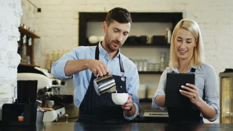 Beautiful-Blonde-Woman-Waitress-Watching-Video-On-The-Tablet-Device-While-Attractive-Waiter-Pouring-Milk-In-The-Cup-Of-Coffee-At-The-Bar