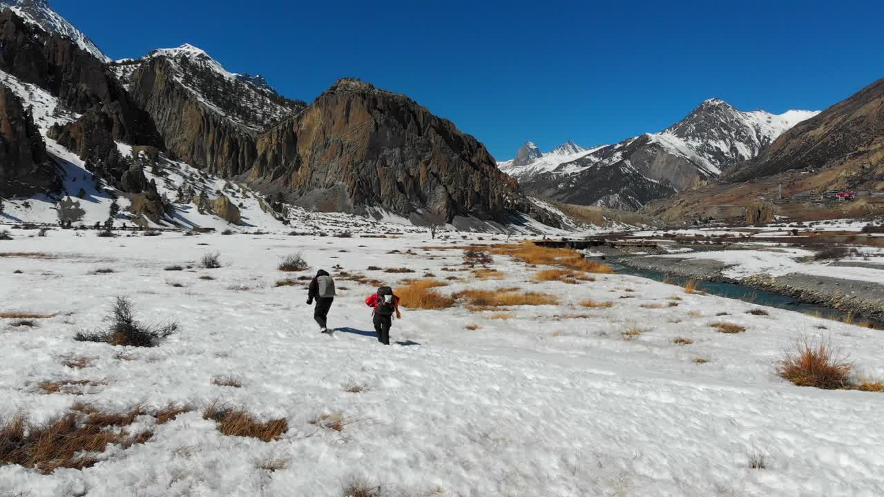 Two travelers trekkers walking hiking through a knee deep snow field. Sunny day in winter. Annapurna Conservation Area, Manang Valley, Nepal. Aerial Drone slider shot passing sliding by.