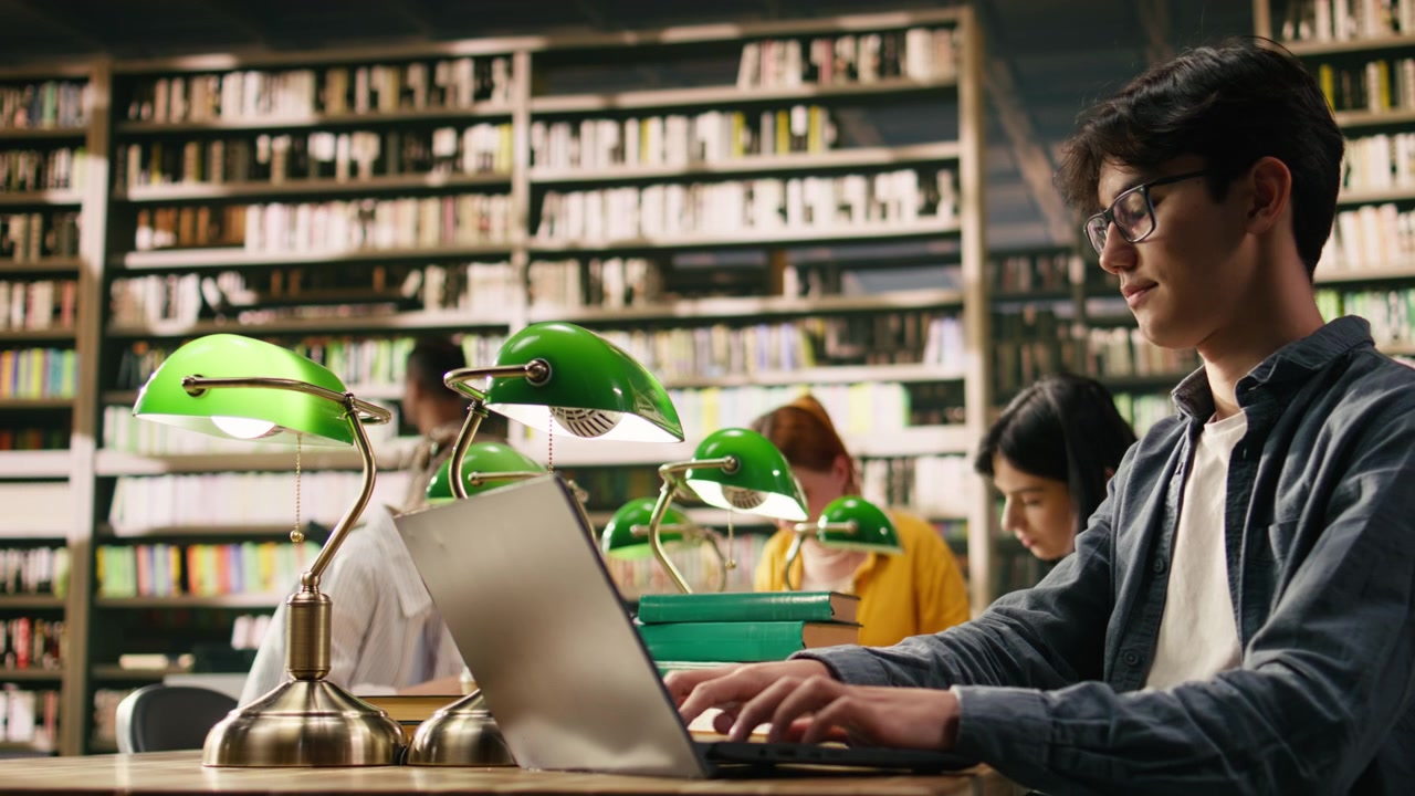 Giovane studente che digita sul laptop in una tranquilla area studio della biblioteca vicino agli scaffali dei libri