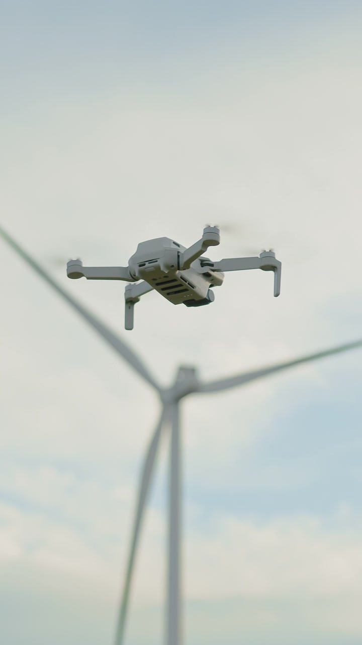 Un piccolo drone bianco si libra nel cielo con una grande turbina eolica sullo sfondo in una giornata nuvolosa