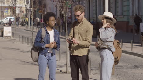 three tourists in the city, two young girls and a young male, checking how a vintage photo camera works