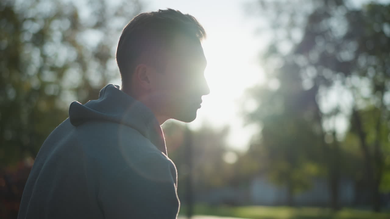Close-up of a guy in gray hoody, facing right with pensive expression partially shaded by sunlight, creating an introspective mood. Background softly blurred with bright daylight highlighting his face