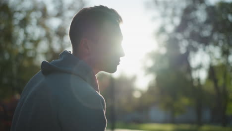 close-up of a guy in gray hoody, facing right with pensive expression partially shaded by sunlight, creating an introspective mood. background softly blurred with bright daylight highlighting his face