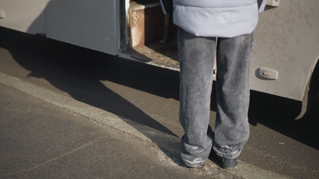 back lower view shows passenger standing beside bus door, waiting to board while others step down, long shadow on curb, worn metal step and asphalt road in daylight, urban commute mood