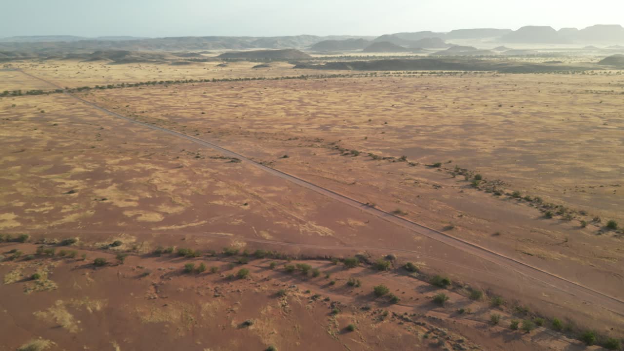 Drooninäkymä Namibiassa: lento punertavien kivivuorten yllä aurinkoisena päivänä. Näkymää ympäröi hiekkasavanni, jossa on joitakin lodgeja ja pysäköityjä autoja; keltaisia ja oransseja sävyjä
