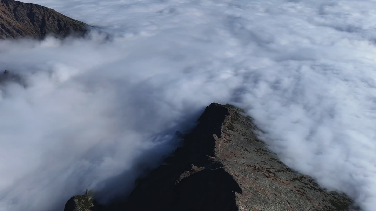Drone-captured aerial top view of a rugged rocky ridge in the High Tatras, partially surrounded by dense clouds. The sharp crest and barren alpine terrain create a dramatic mountain scene