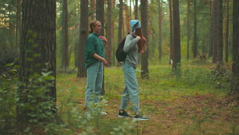 young woman in green assists her friend in putting on a backpack in a tranquil forest setting with a warm glow of sunlight filtering through trees, they look at each other and walk together