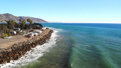 aerial drone shot rising up over ocean waves crashing on the rocky coast with palm trees and rv campground on a california beach