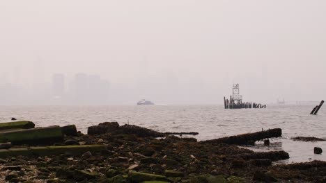 View-of-Manhattan-covered-in-smoke-from-wildfires-seen-from-beach-on-the-east-river-with-waves-crashing-on-mossy-rocks-in-the-foreground-and-ferry-crossing-river