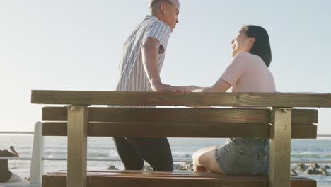 happy biracial couple sitting on bench on promenade, in slow motion