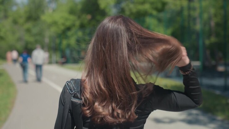 Primo piano di una donna con lunghi capelli castani che cammina all'aperto su un sentiero, indossa un top nero e porta uno zaino. I capelli si muovono naturalmente mentre la donna cammina in un ambiente simile a un parco con alberi verdi.