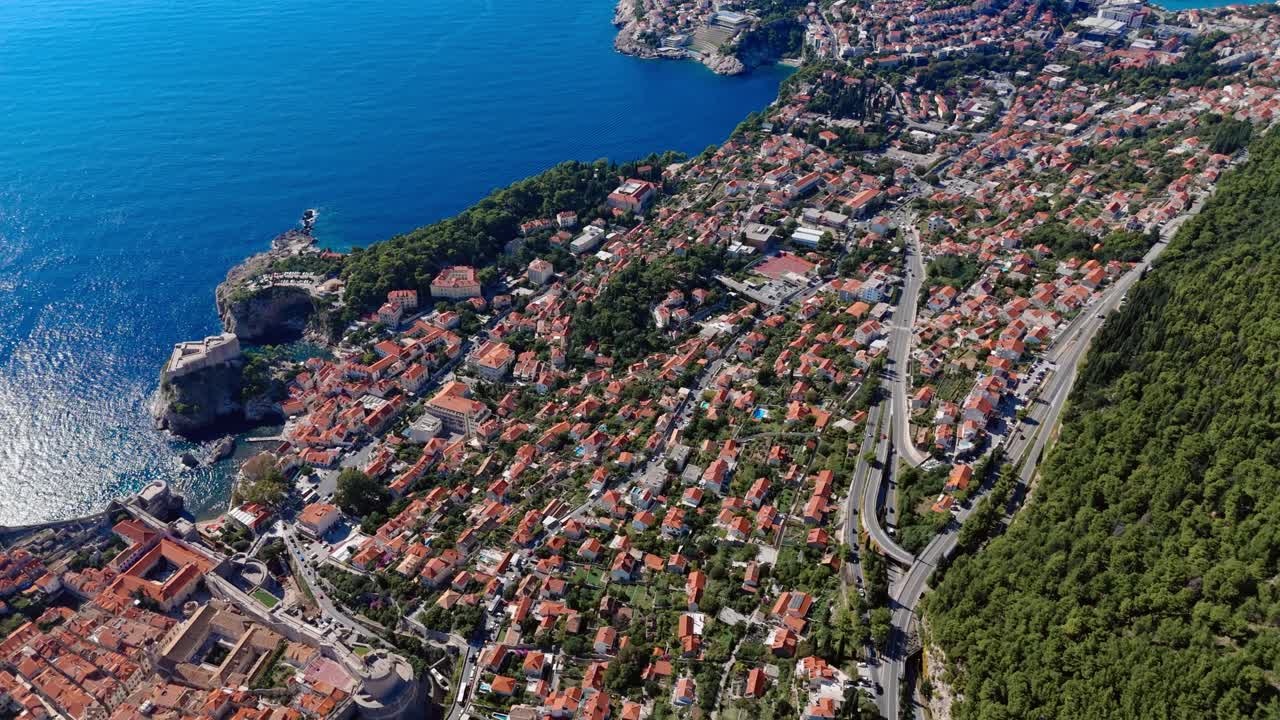 Aerial landscape drone flies over Dubrovnik near the Old Town, capturing terracotta rooftops, the deep blue Adriatic Sea, and rocky coastline bathed in bright sunlight along the Mediterranean shore
