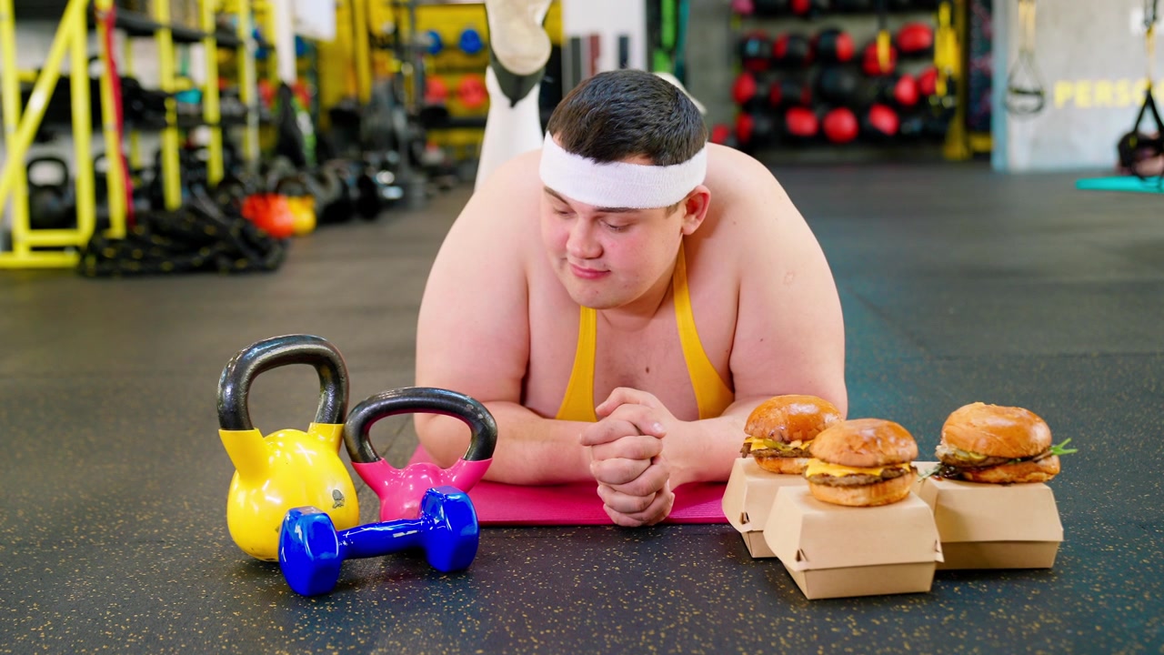 Uomo in sovrappeso che contempla hamburger e attrezzatura da palestra
